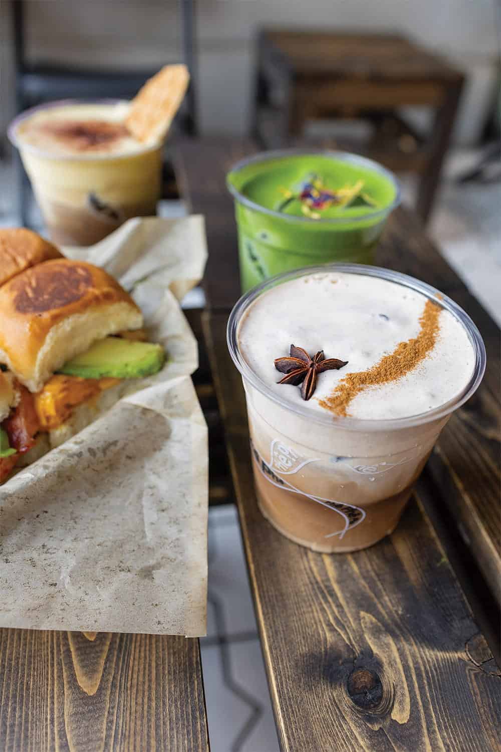 A cold coffee drink featuring Vietnamese coffee flavors is garnished with star anise and cinnamon, sitting on a wooden table alongside a sandwich, matcha drink, and another beverage in the background.