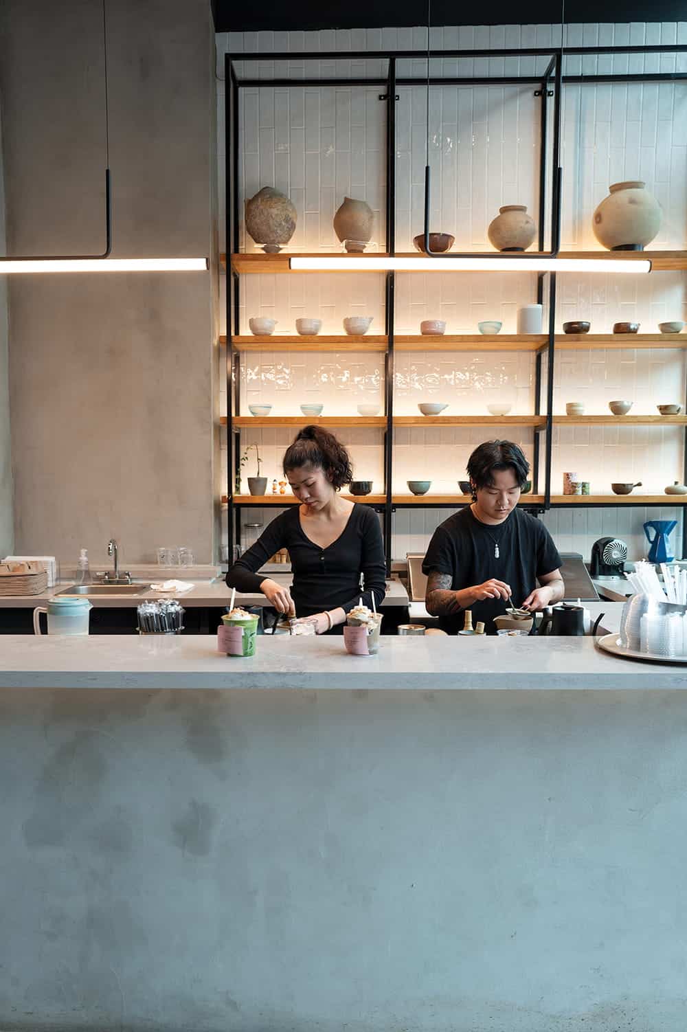 Two people are preparing drinks behind a modern coffee bar, where shelves of cups and pottery nod to Vietnamese coffee traditions, creating a unique atmosphere among coffee shops in Seattle.