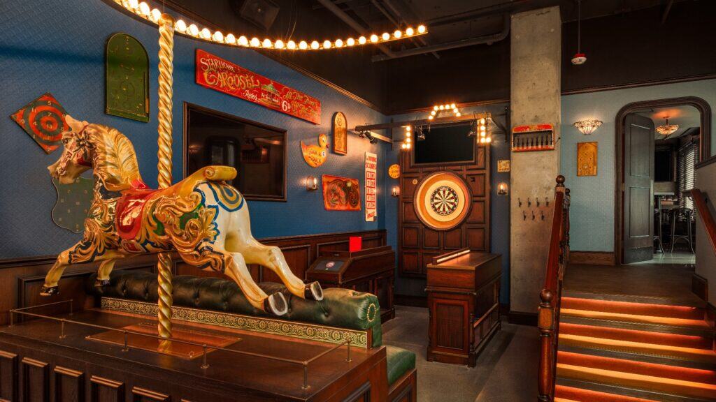 A vintage-themed room features a decorative carousel horse, dartboard, carnival signs, wood paneling, and orange-lit stairs.