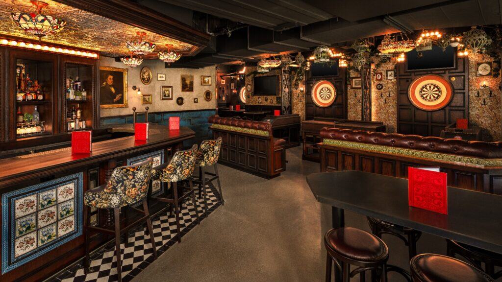 A cozy pub interior with a bar, patterned stools, leather sofas, dartboards, vintage décor, framed art, and red menus on tables.