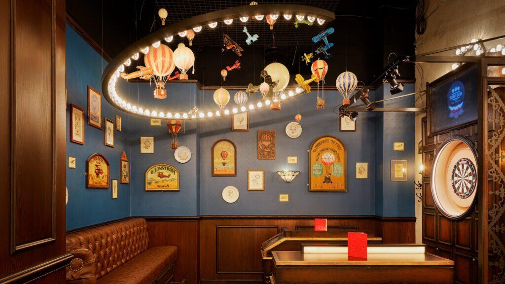 A cozy game room with a dartboard, brown leather seating, blue walls decorated with framed art, and a ceiling display of hot air balloons and model airplanes under ring lights.