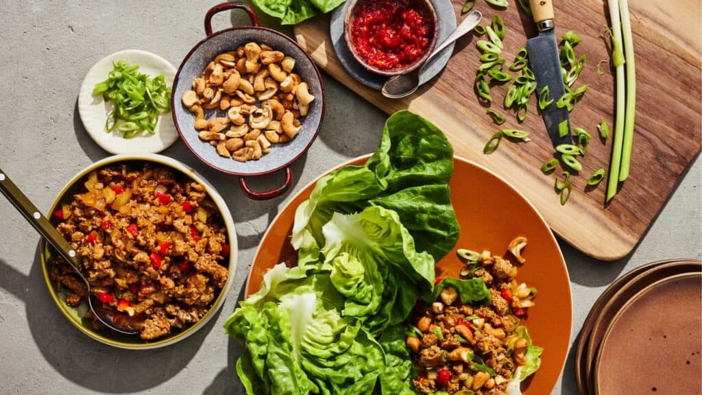 A table with lettuce leaves, seasoned ground meat with vegetables, cashews, sliced green onions, sauce, and a cutting board with a knife and more green onions—perfect for anyone seeking tasty high protein recipes.