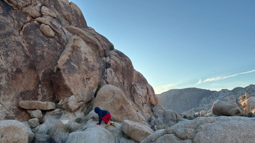 A person wearing a hoodie and red pants climbs over large rocks at the base of a rocky mountain under a clear sky.