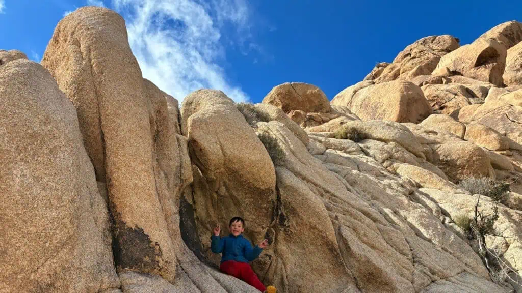 A child sits among large tan rock formations under a blue sky with some clouds, wearing a blue jacket and red pants.