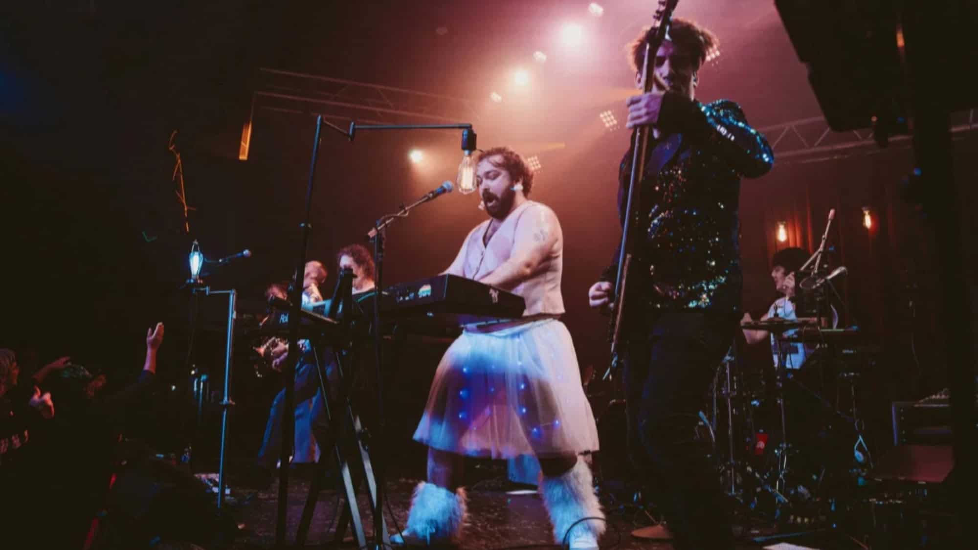 A band performs on stage under colorful lights; the keyboardist is wearing a white tutu and furry boots, while other musicians play guitar and drums.