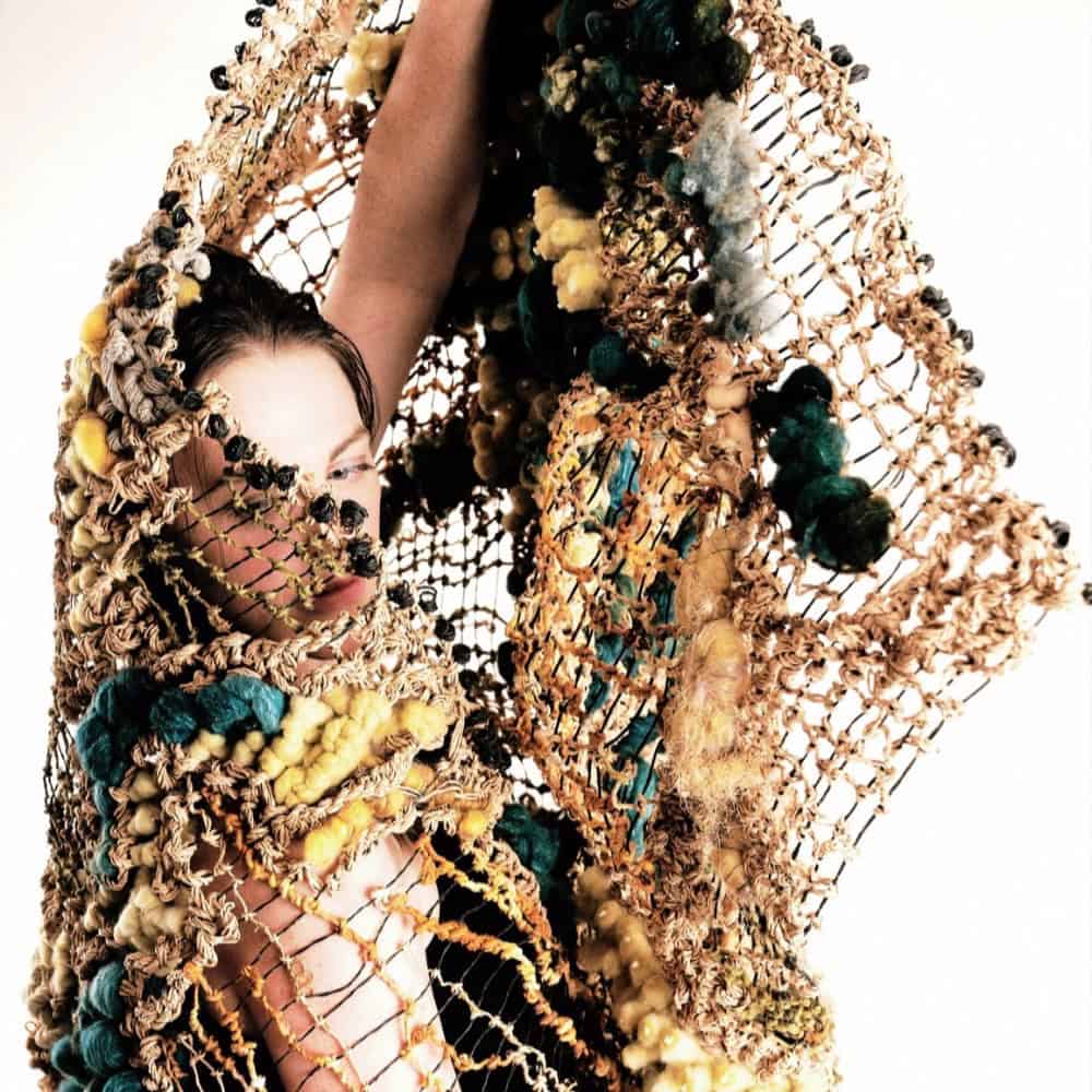 A person partially covered by a large, textured, woven net with blue, yellow, and brown yarn details, set against a plain background.