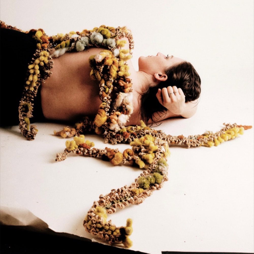 A person lies on their side with their back exposed, draped in long, textured strands of multicolored material resembling yarn or organic matter, against a plain background.