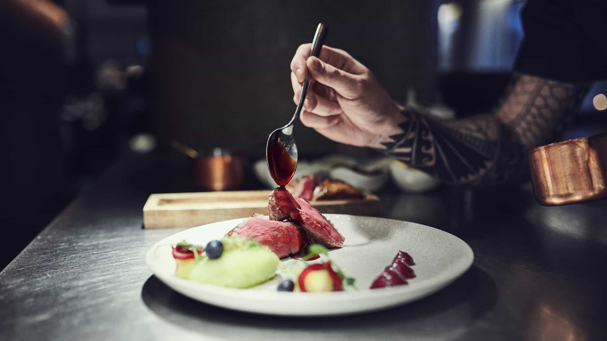 A chef with a tattooed arm drizzles sauce over slices of meat on a white plate, garnished with vegetables and berries, in a professional kitchen.