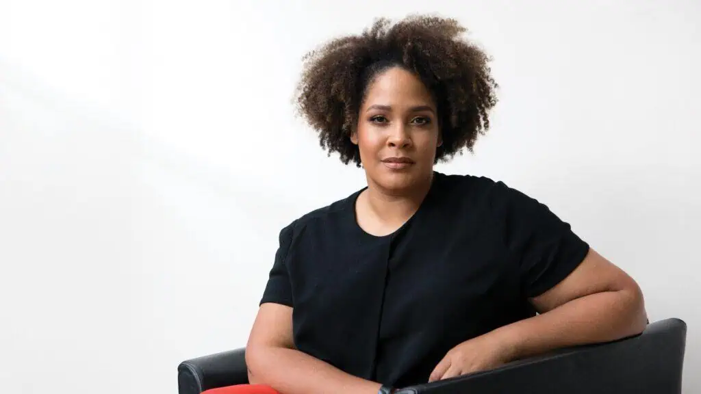 A person with curly hair sits in a black chair against a plain white background, wearing a black shirt and red pants, looking directly at the camera.