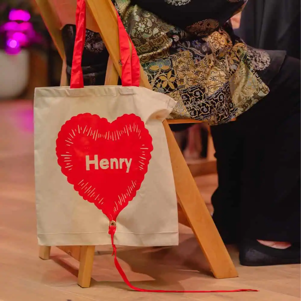 A beige tote bag with red handles hangs from a chair. The bag features a large red heart graphic with the name "Henry" printed in the center.