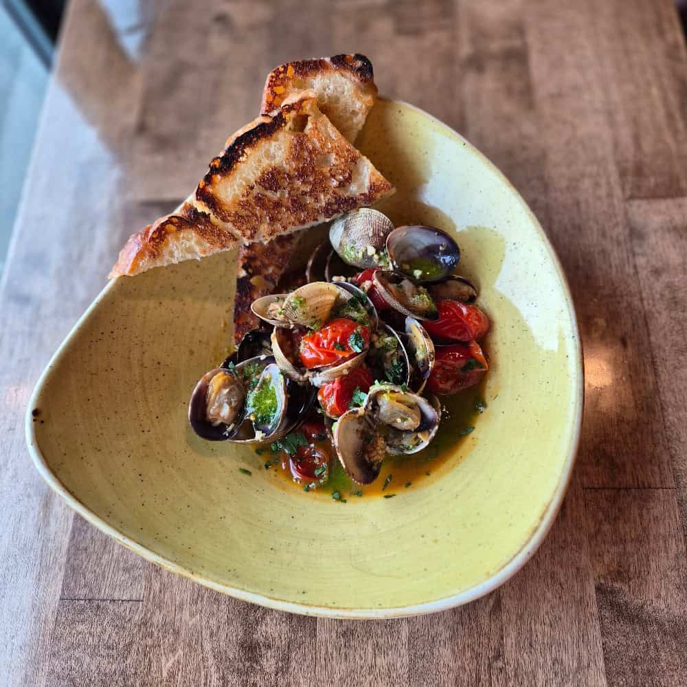 A bowl of steamed clams with cherry tomatoes and herbs, served with two slices of toasted bread on a yellow oval plate on a wooden table.
