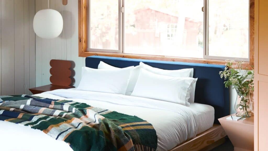 A neatly made bed with white linens and a green patterned blanket is positioned under a window in a bright, modern bedroom with wood accents and a bedside table holding a vase of flowers.