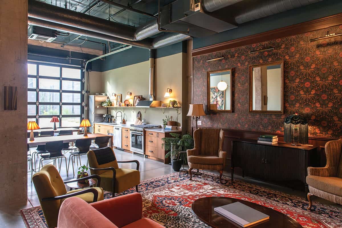 Spacious living area with vintage furniture, patterned rug, open kitchen, dining table, and large industrial windows letting in natural light—a perfect space for gathering and enjoying life Better Together.