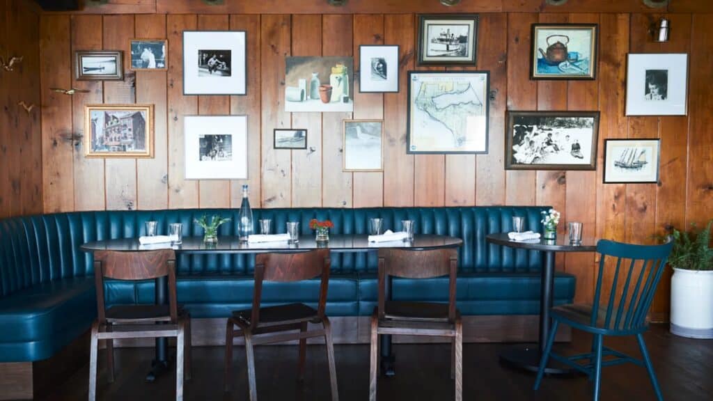 Restaurant seating area with dark wood chairs, blue tufted booth, tables set for dining, and a wood-paneled wall decorated with framed artwork and photographs.