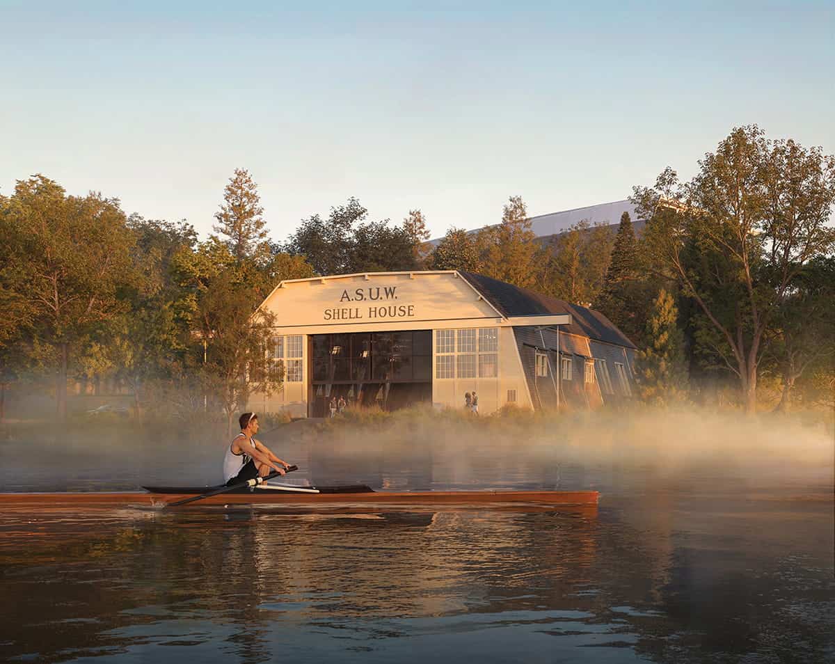 A person rows a single scull on a calm, misty lake near the A.S.U.W. Shell House, surrounded by trees—a striking scene reflecting design firm Mithun’s appreciation for harmony between architecture and nature.