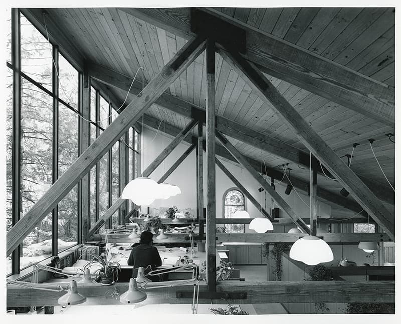 Black and white photo of Mithun’s office, featuring exposed wooden beams, large windows, multiple desks, and people working under hanging lights.