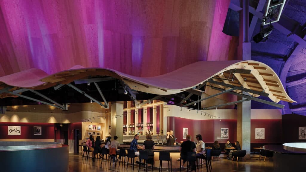 People sit at a modern bar with a wavy wooden ceiling and purple lighting, surrounded by art on dark red and beige walls, showcasing the creative vision of design firm Mithun.