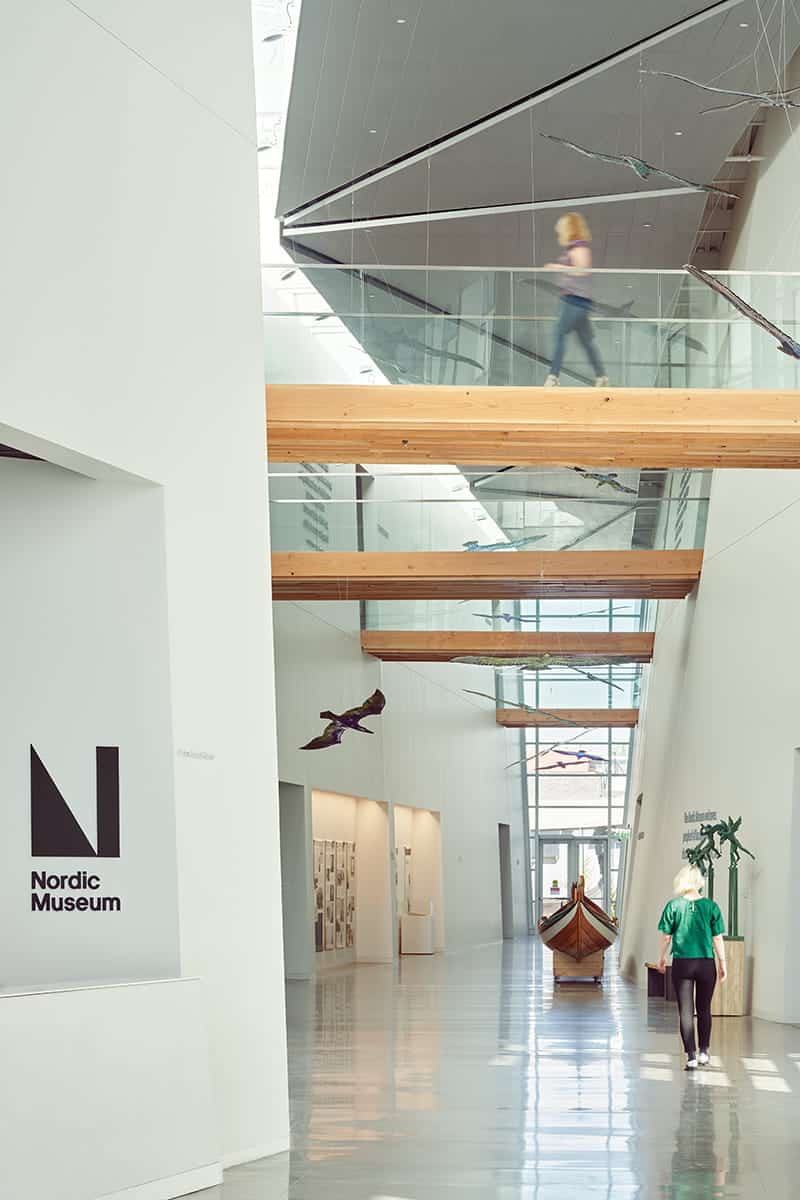 Interior of the Nordic Museum, designed by Mithun, features a wooden boat, hanging bird sculptures, two people walking, and large windows letting in natural light.