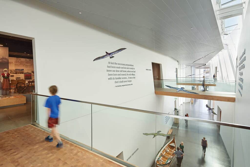 A modern museum interior by design firm Mithun, with people walking, features suspended model sea creatures and text on the wall above a glass railing and wooden floor.
