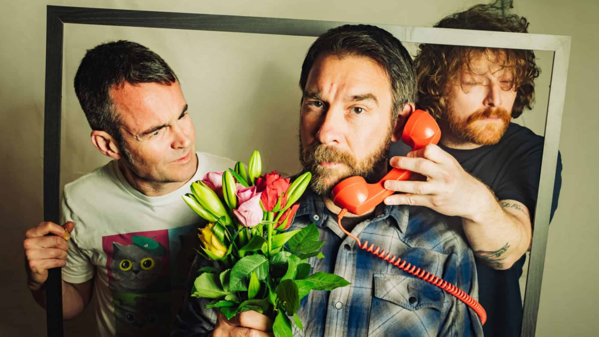 Three men pose inside a rectangular frame; one holds flowers, another holds a red telephone to his ear, and the third stands behind them, looking at the man with flowers.