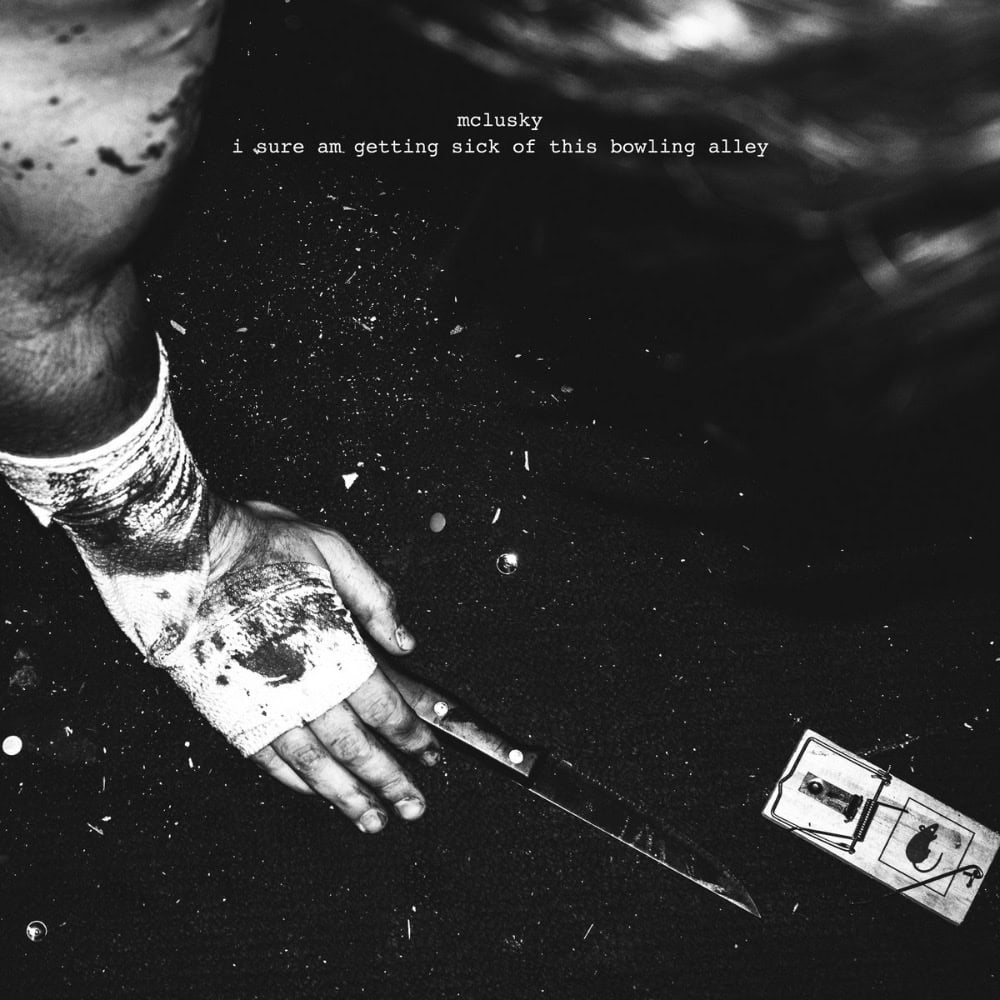 A bandaged, bloodied arm reaches for a knife on a gritty surface near a mousetrap; text above reads "mclusky i sure am getting sick of this bowling alley.