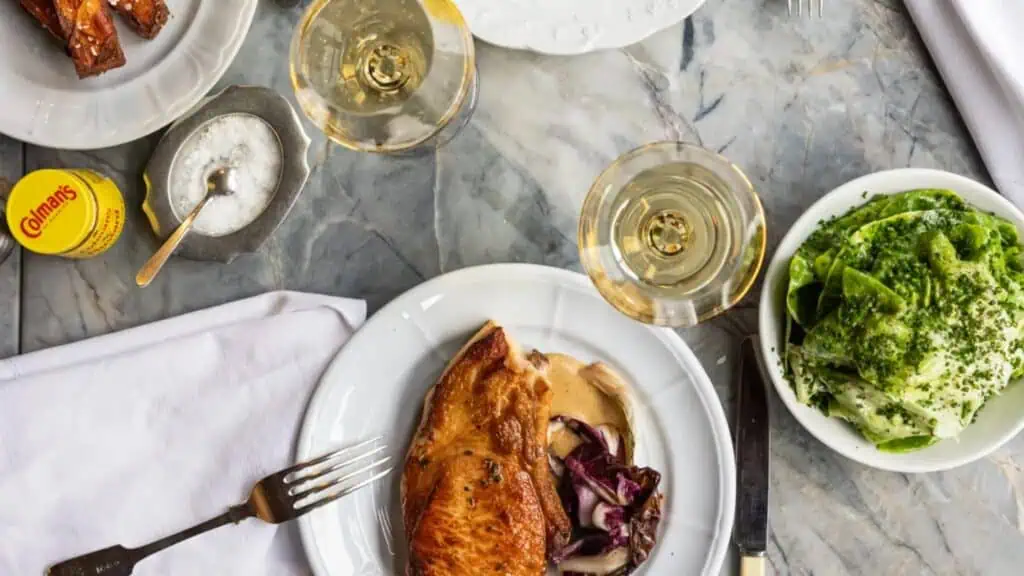 A plate with a piece of roasted chicken, sauce, and red cabbage—reminiscent of traditional dishes—next to a bowl of green salad, two glasses of white wine, salt, and mustard on a marble table.