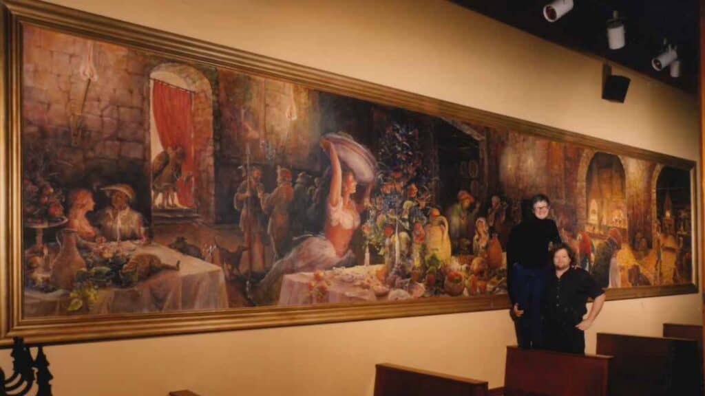 Two people stand in front of a large, detailed mural depicting a festive banquet scene with various figures, food, and activities in a warm, indoor setting.