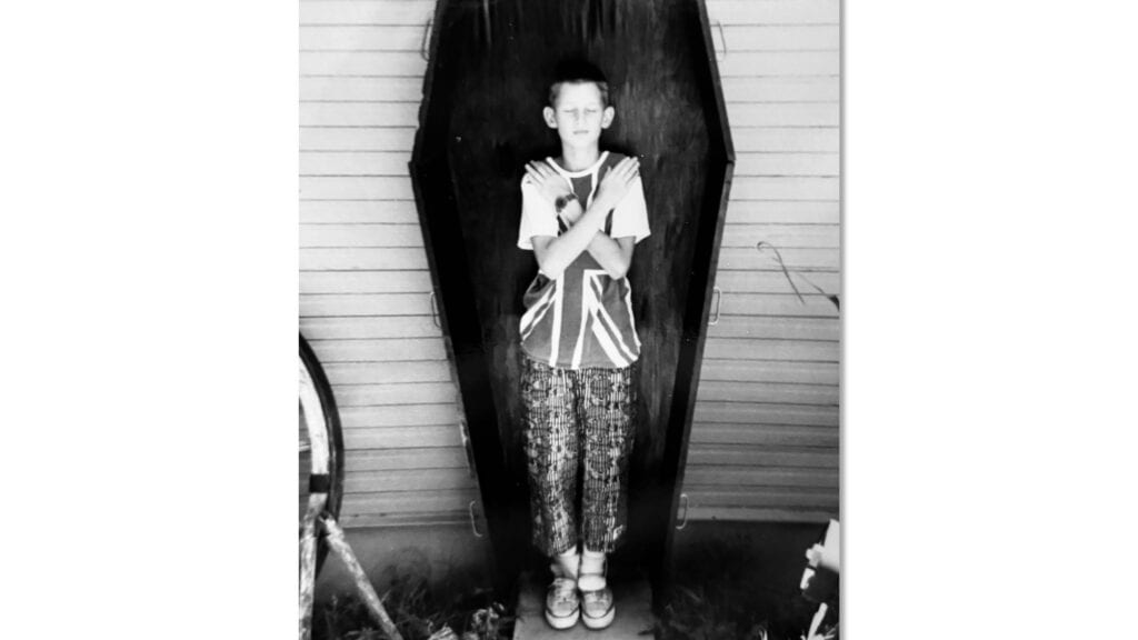 A boy stands inside an upright coffin with his arms crossed over his chest, in front of a wooden wall.