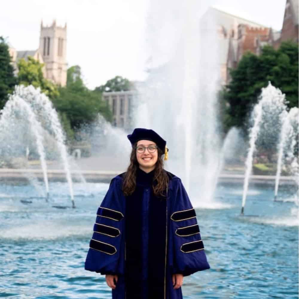 A person in academic regalia stands smiling in front of a large fountain with university buildings and trees in the background.