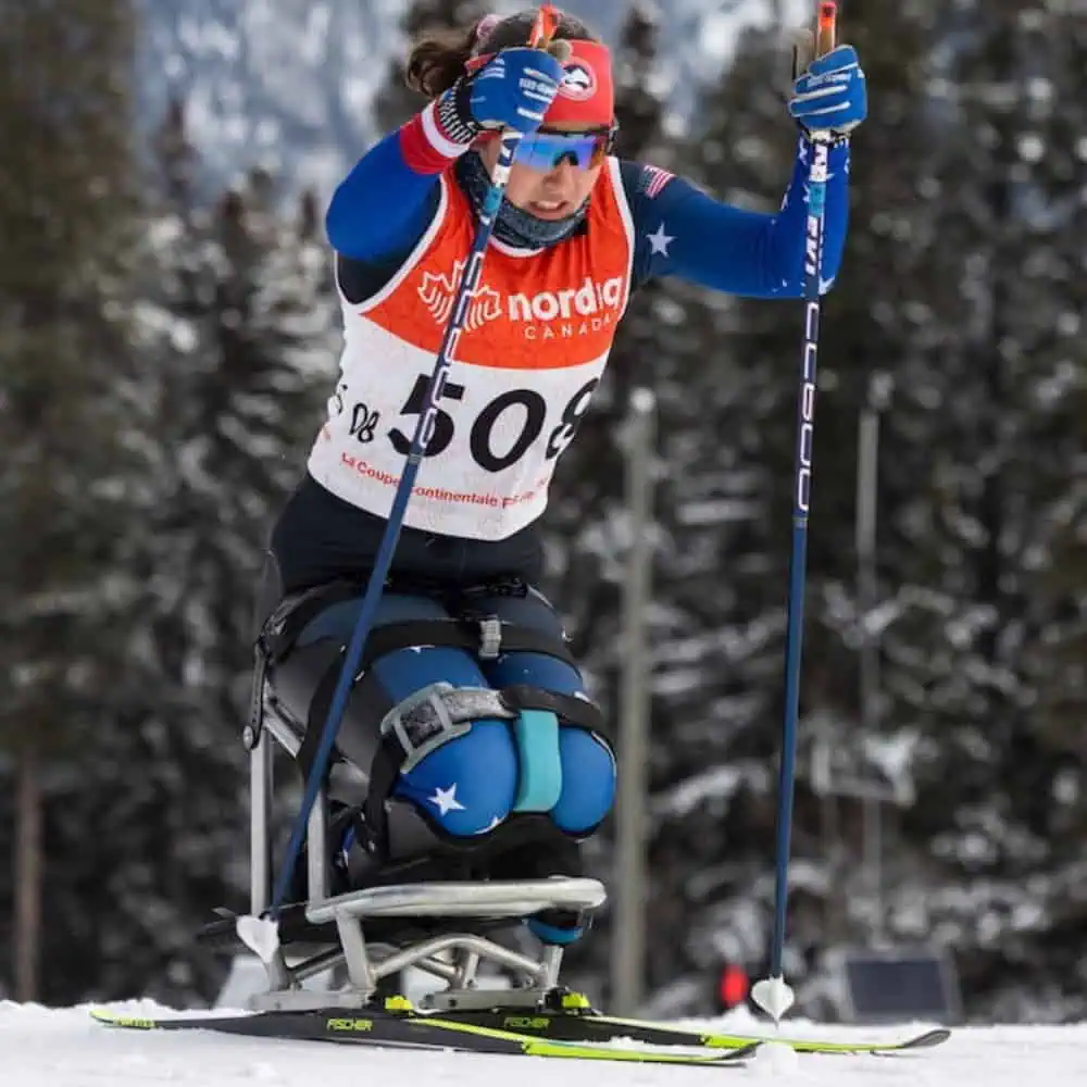 An athlete using a sit-ski and ski poles competes in a cross-country skiing event on a snowy course, wearing a race bib and winter gear.