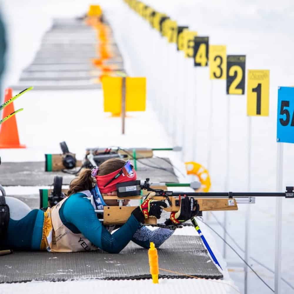 A biathlete in winter gear lies prone, aiming a rifle at numbered targets during a snow-covered biathlon shooting event.