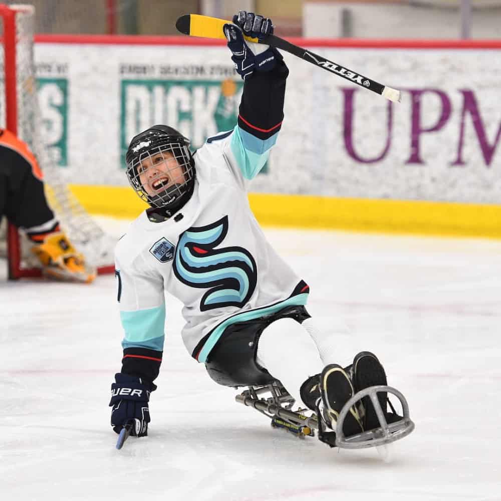 A sled hockey player in a white jersey celebrates on the ice with one arm raised, holding a hockey stick.