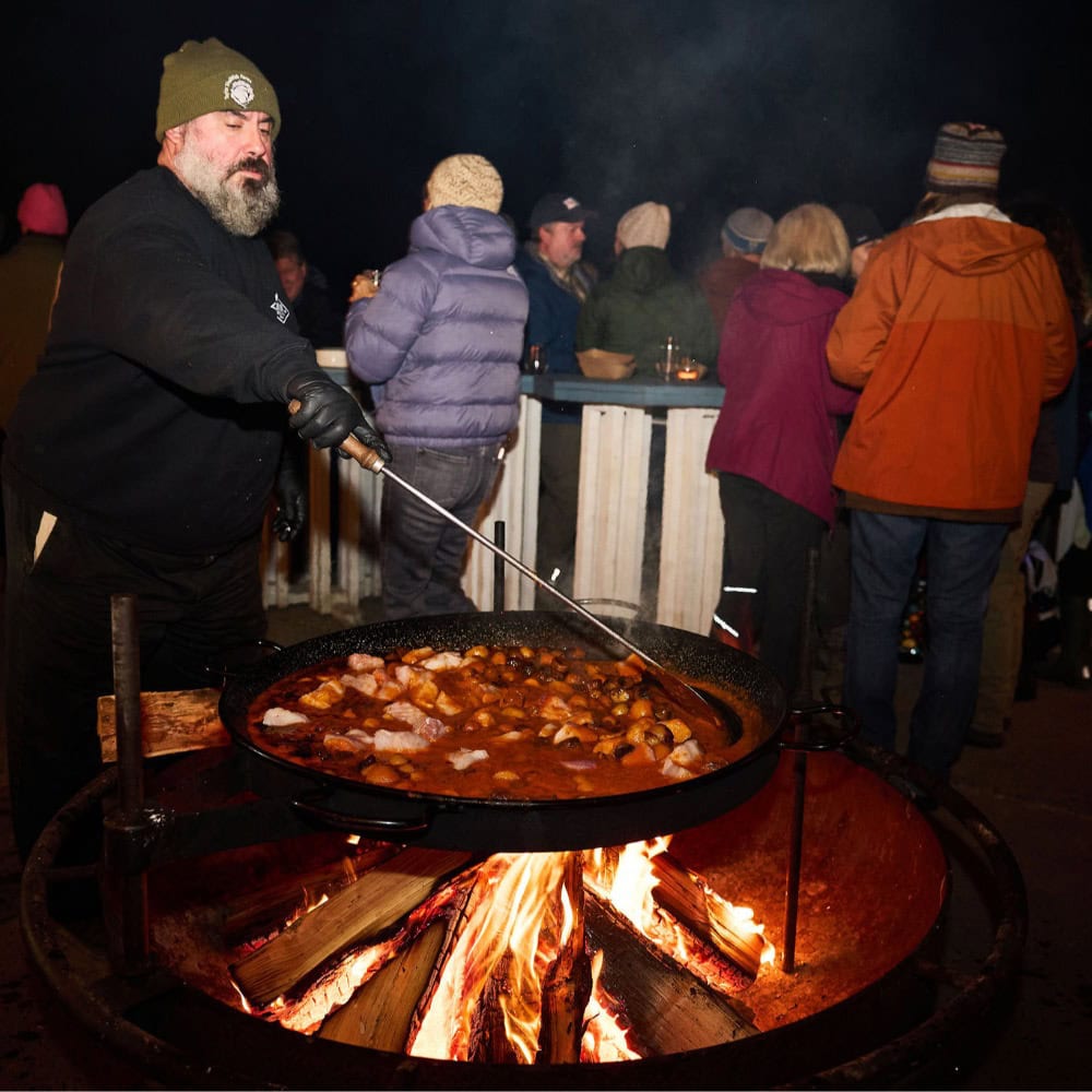 A man cooks a large pan of food over an open fire outdoors at night, while a group of people in winter clothing gather in the background.