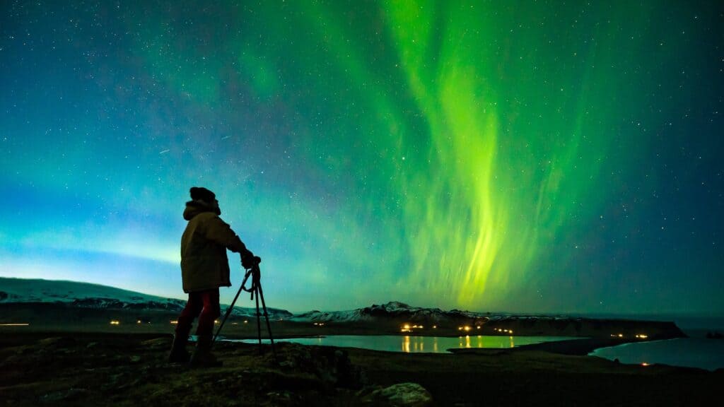 A person stands with a tripod, watching green aurora borealis lights in the night sky over a landscape with water and distant mountains.