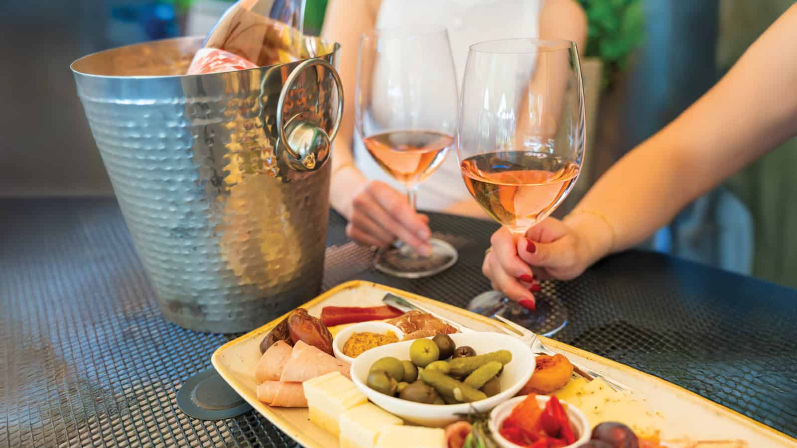 Two people holding glasses of rosé wine sit at a table with a platter of cheese, olives, pickles, and meats, enjoying the vibrant winery scene that makes Woodinville one of the best places to live in Washington. A metal ice bucket chills bottles nearby.