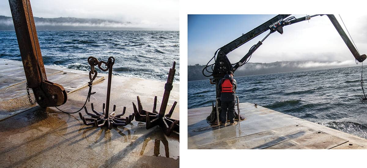 Two metal anchors rest on a wet dock by the water near Hood Canal; a person in a life jacket operates a crane at the dock’s edge, where oysters are harvested in the misty Pacific Northwest.