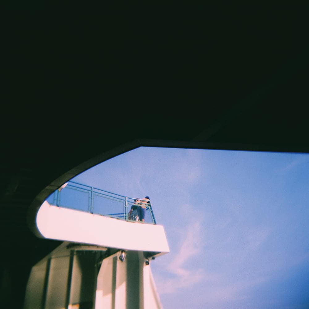 A person stands alone on the upper deck of a ship, looking out over the railing under a blue sky. The image, part of a Ferry Therapy photo essay, is partially framed by a large dark shadow.
