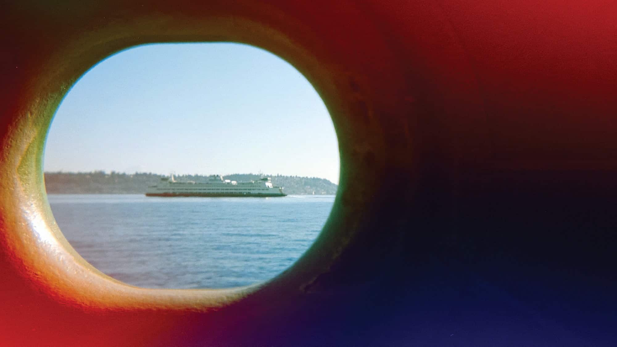 A ferry on the water is seen through a round window, with land and trees in the background and red light leaks framing the image—a serene moment perfect for any photo essay capturing subtle ferry therapy.