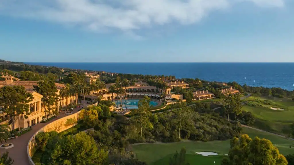 Aerial view of a coastal luxury resort with a pool, lush landscaping, and golf course, overlooking the ocean under a partly cloudy sky.