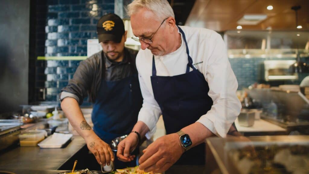 Two chefs wearing aprons prepare food in a restaurant kitchen, focusing on plating a dish together under bright lighting—like actors collaborating at Seattle Rep, their teamwork reflected as vividly as memories in a rearview mirror.