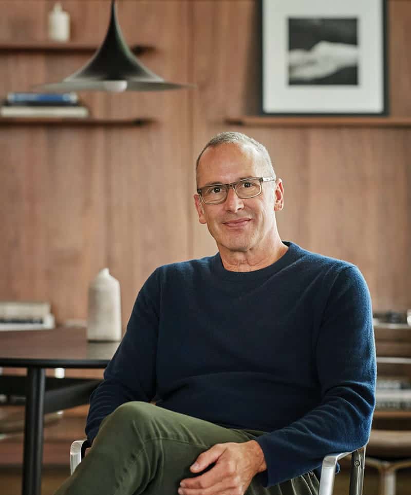 A man with glasses and a blue sweater sits in a modern room, surrounded by wooden walls, shelves, and framed artwork—an environment that reflects an appreciation for craft and value.