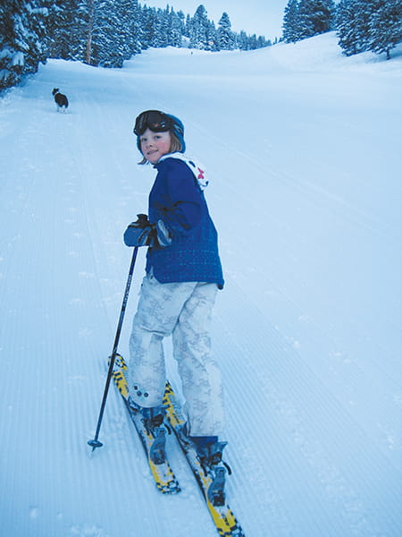 A child in winter gear stands on skis, looking back while holding ski poles on a groomed snowy slope—channeling skimo spirit like professional runner Anna Gibson—with trees and a dog visible in the distance.