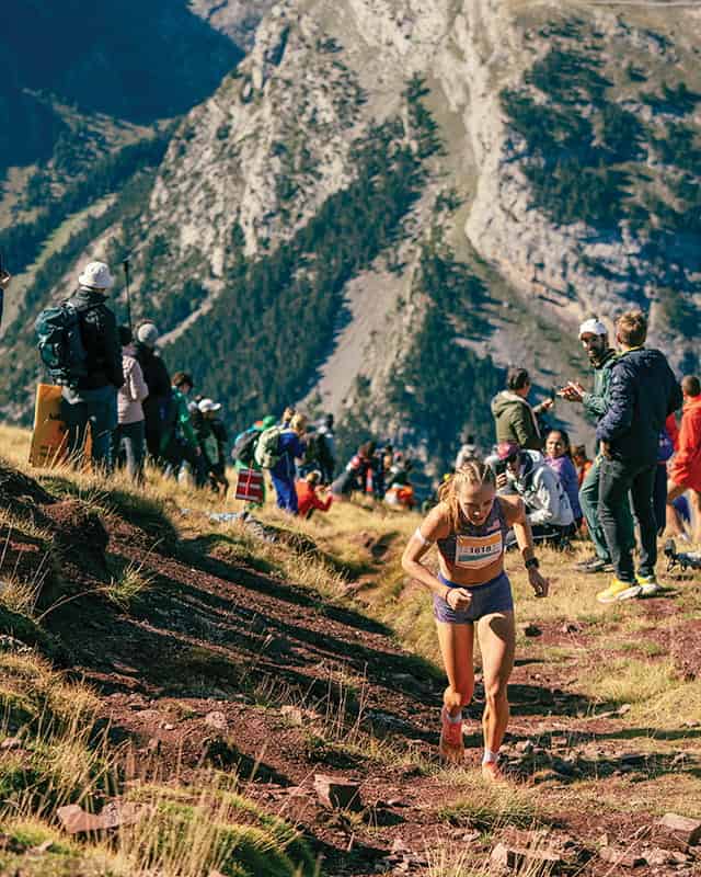 Professional runner Anna Gibson competes in a mountain trail race as spectators watch from the side, with rugged mountains visible in the background.