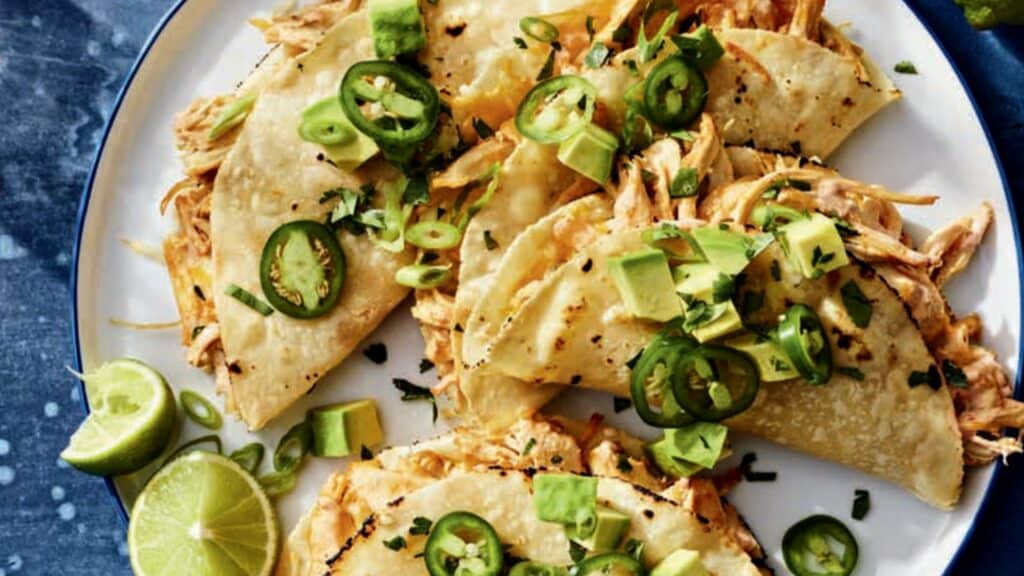 Three shredded chicken tacos topped with sliced jalapeños, avocado, green onions, and cilantro, served with lime wedges—a flavorful dish inspired by high protein recipes from top cookbooks.
