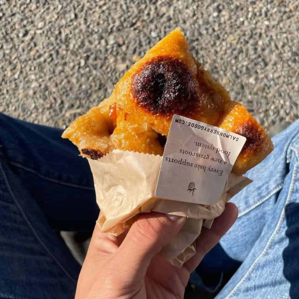 A hand holds a piece of browned bread wrapped in paper, with a label on top—a perfect March food pick. The person is sitting outdoors on a gravel surface, wearing blue jeans.