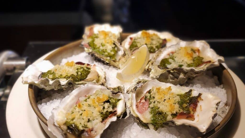 A plate of baked oysters topped with herbs and breadcrumbs is served on a bed of coarse salt with a lemon wedge in the center.