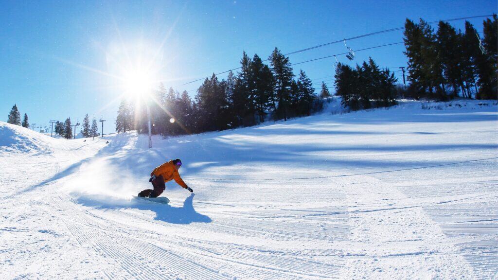 A snowboarder in an orange jacket carves down a groomed, snowy slope under a clear blue sky with trees and ski lift cables in the background.