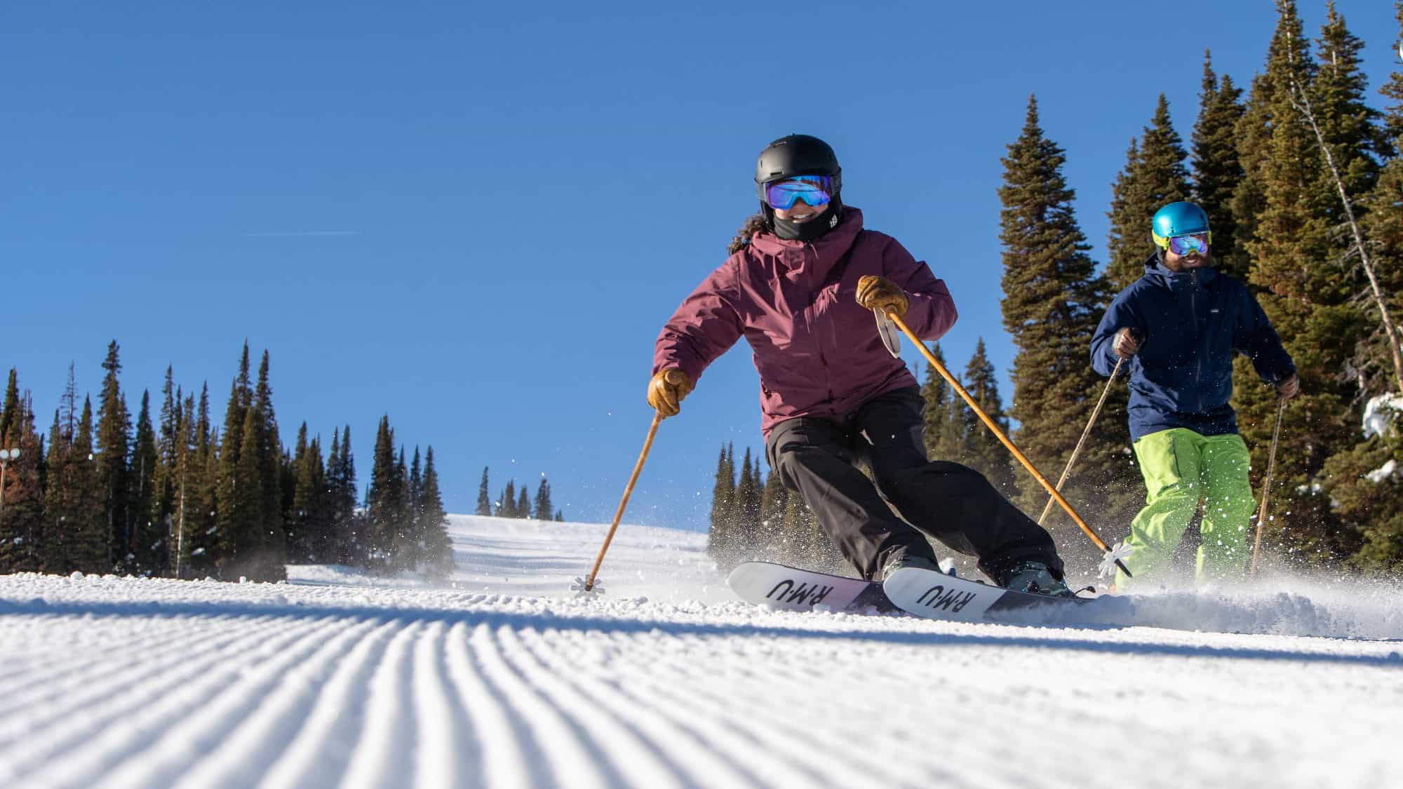 Two people ski downhill on a groomed snow slope with pine trees and a clear blue sky in the background.