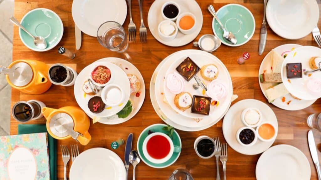 A wooden table set for afternoon tea with plates of pastries, sandwiches, scones, teapots, cups, cutlery, and jars of jam and cream arranged neatly.