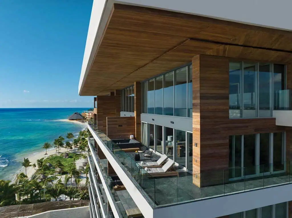 Modern beachfront condominium with glass balconies and wood paneling overlooks a sandy beach, palm trees, and clear blue ocean—perfect for relaxation and wellness in Mexico under a sunny sky.